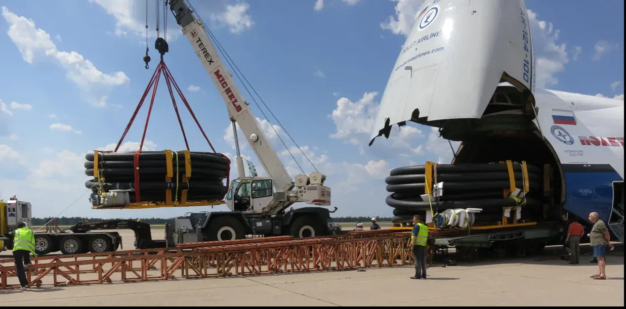 Crane lifting a coiled pipe carousel on the apron beside a cargo aircraft with its nose raised