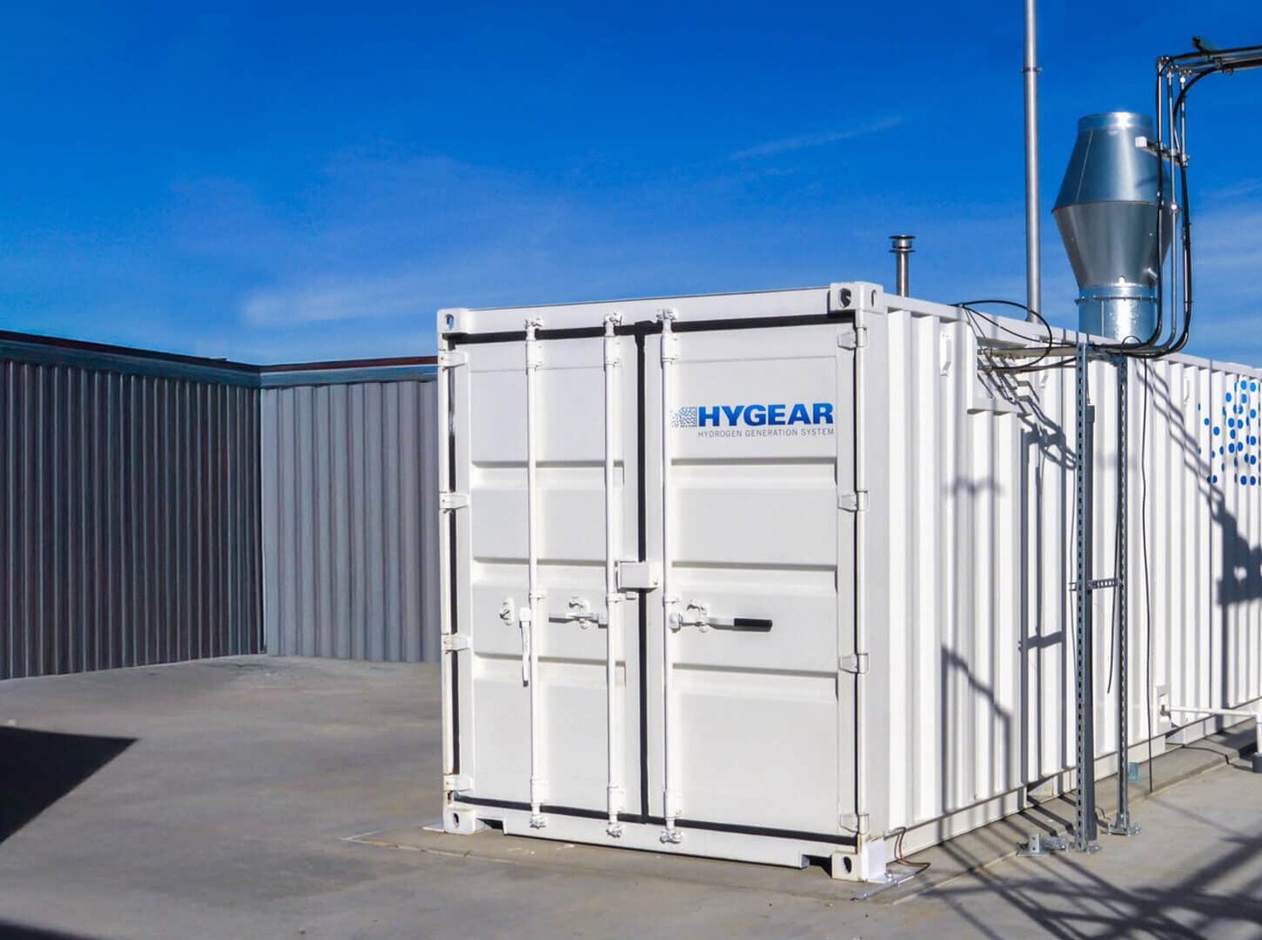 A white containerised HyGear Hy.GEN hydrogen generation system installed outdoors under a clear blue sky, with an exhaust stack and connected pipework alongside
