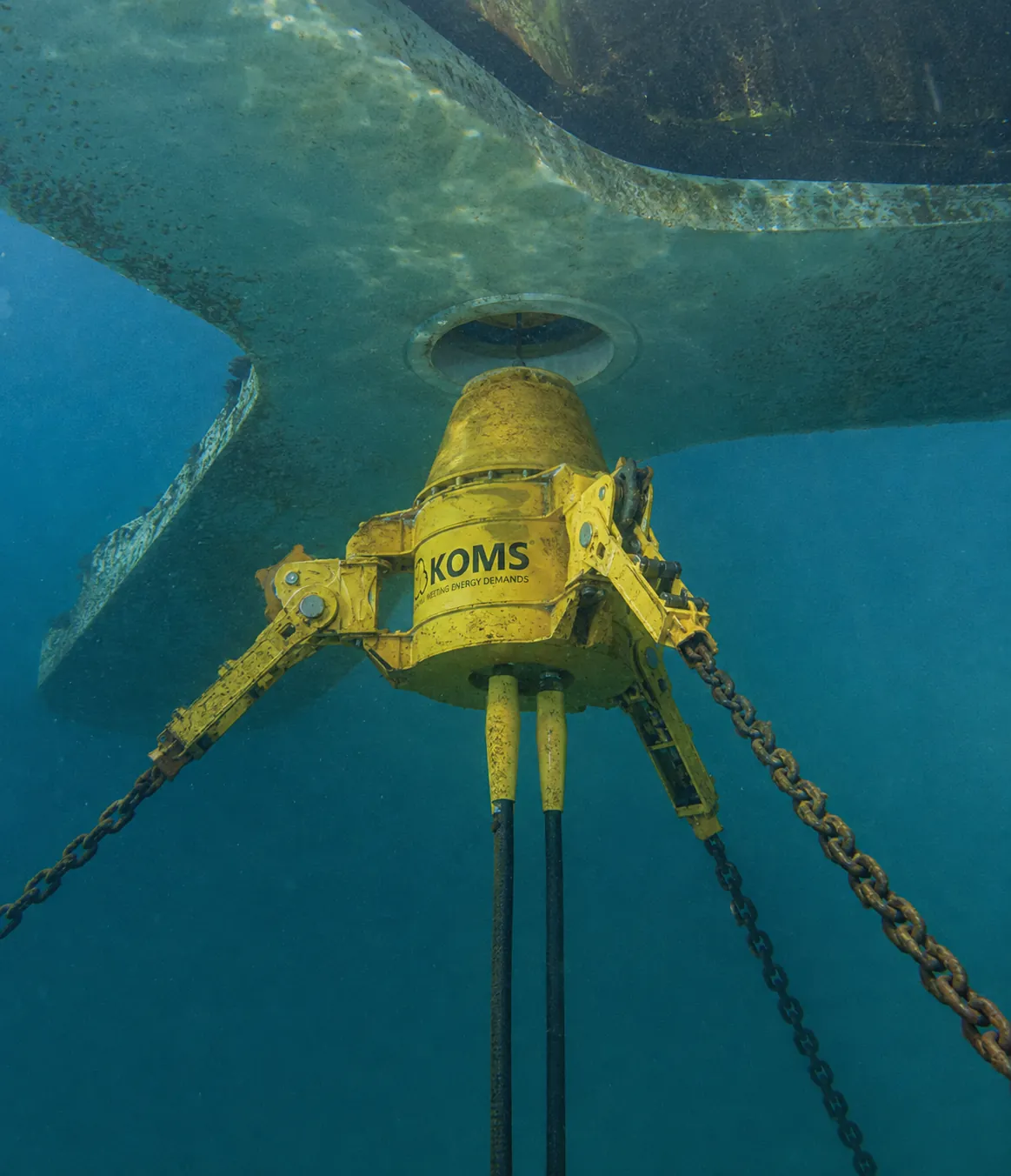 A yellow subsea quick release mooring connector seen underwater beneath the hull of a floating platform, with heavy mooring chains attached to each arm